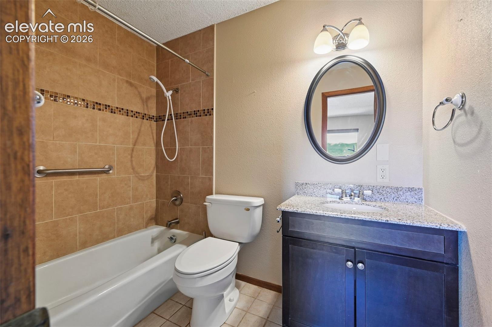 Bathroom featuring a dark-finish vanity with a stone-composite countertop, shower-tub combination with tile surround, oval wall-mounted mirror, and polished-finish hardware