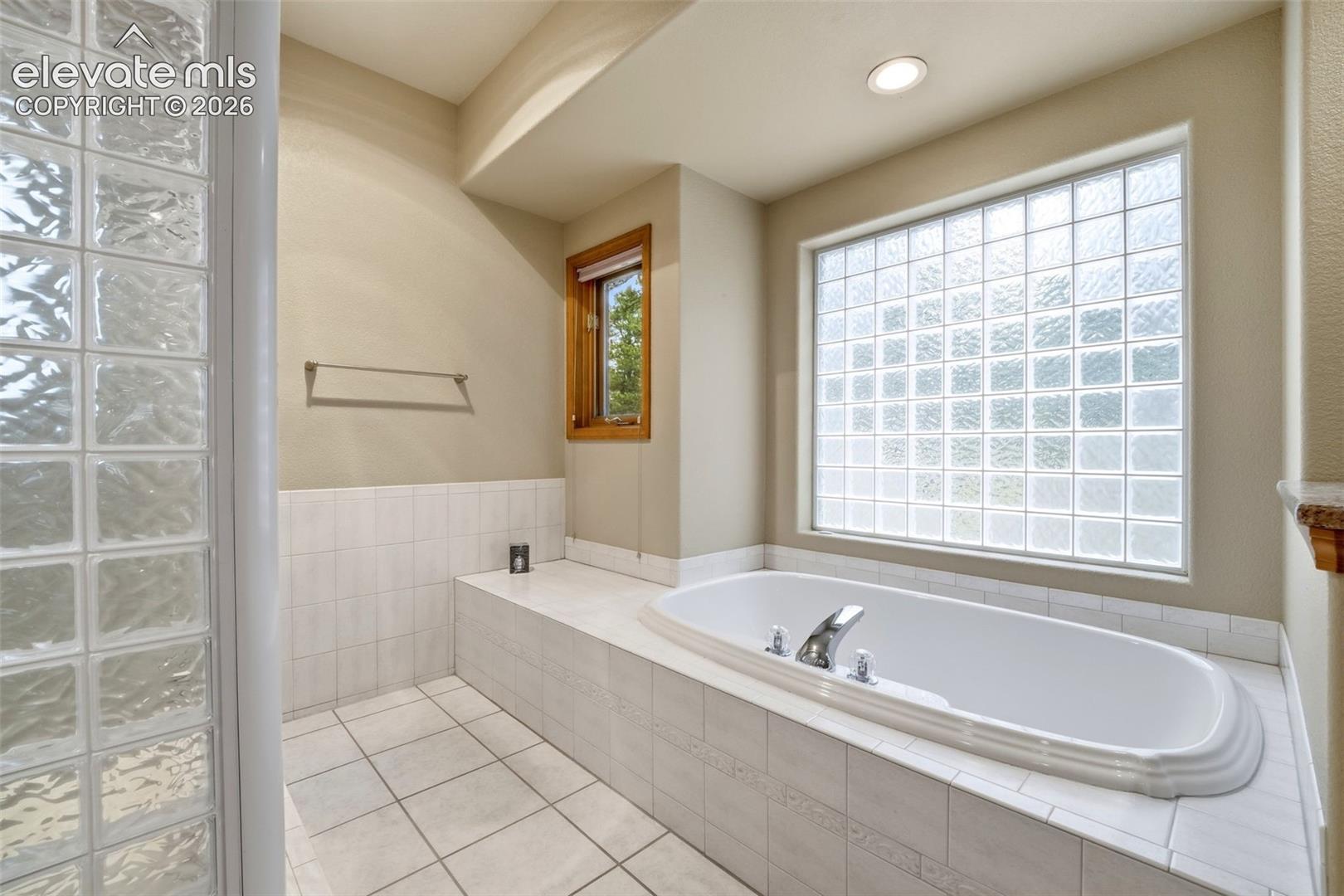 Bathroom featuring a built-in soaking tub with chrome fixtures, a large glass block window, and white tile flooring