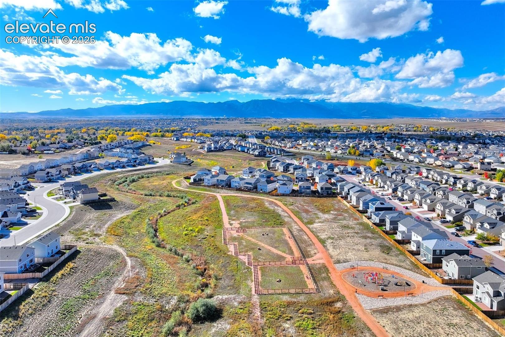 Aerial perspective of expansive open space featuring natural terrain, winding pedestrian paths, and a developed playground area against a backdrop of mountain ranges