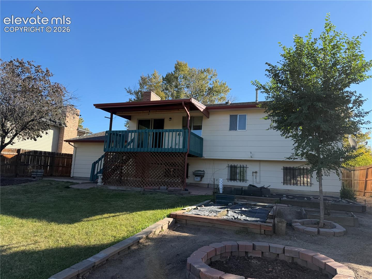 Rear exterior featuring a raised wooden deck with lattice skirting, brick chimney, wood-paneled siding, and a backyard with a stone-bordered fire pit and planter boxes
