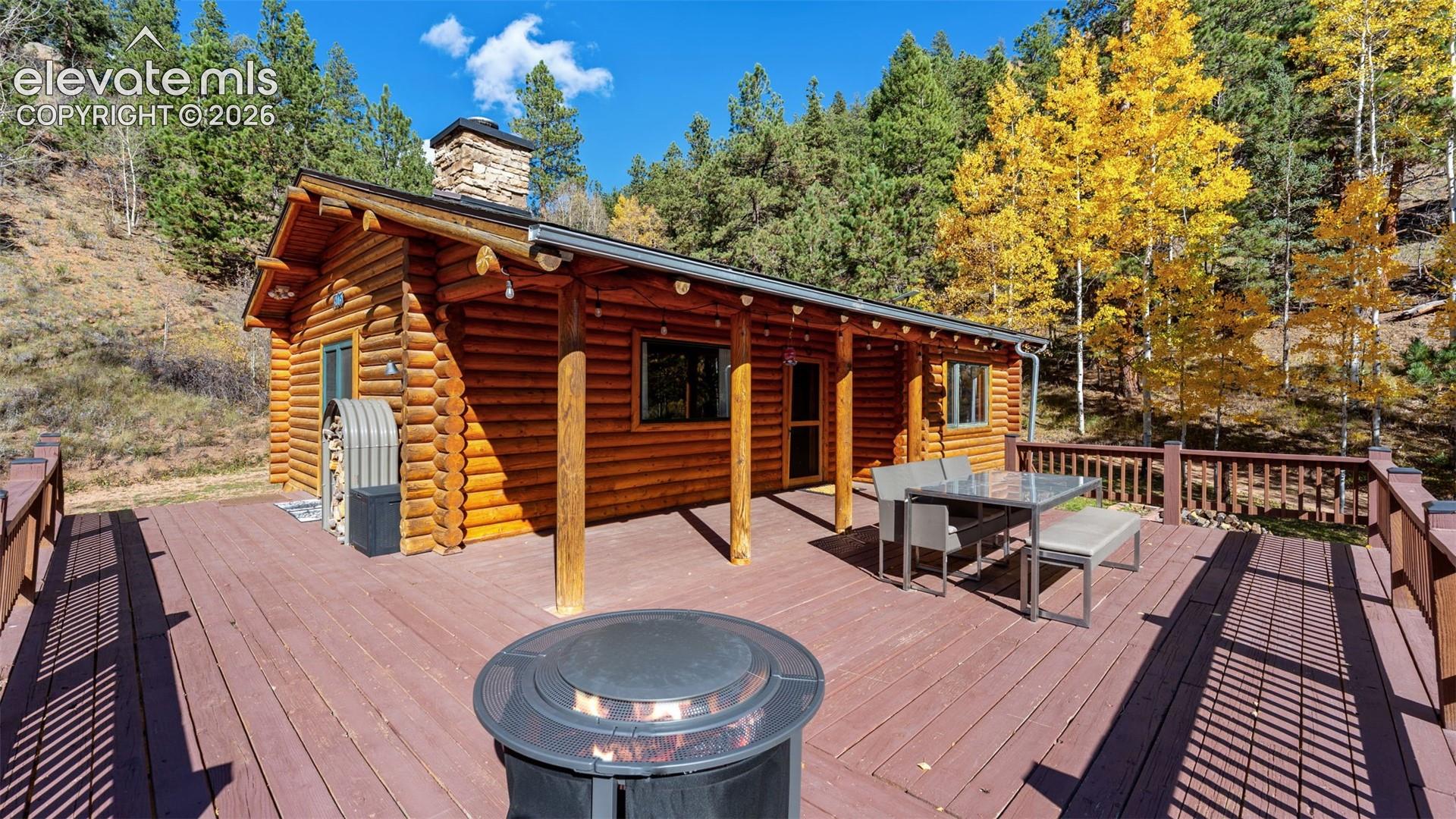 Log exterior cabin featuring a covered porch, stone chimney, and expansive wood-finish deck with integrated railing