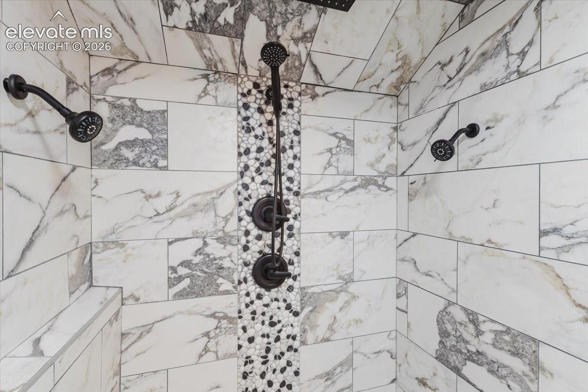 Shower enclosure featuring white marble-look tile with gray veining, dark bronze fixtures, a multi-function shower head, a handheld shower spray, and a pebble tile accent strip