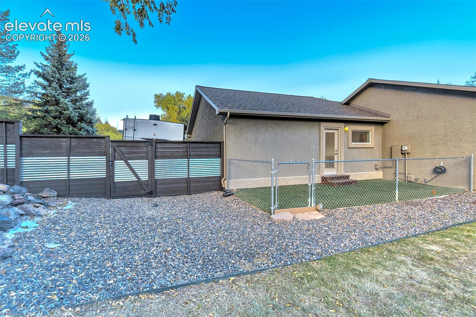 Exterior of stucco home featuring a fenced artificial turf area, decorative horizontal privacy fencing with metal corrugated paneling, and stone ground cover