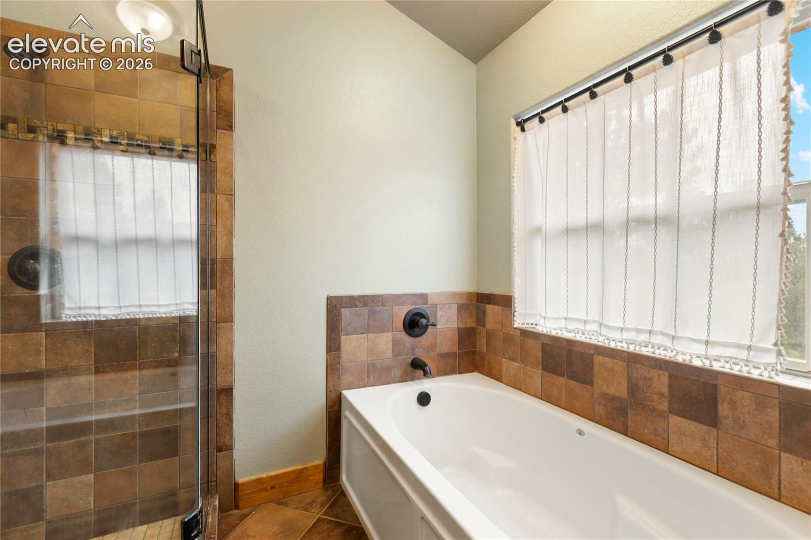 Bathroom featuring a built-in soaking tub, a tiled walk-in shower with a glass enclosure, square earth-tone wall tiling, and matte black plumbing fixtures