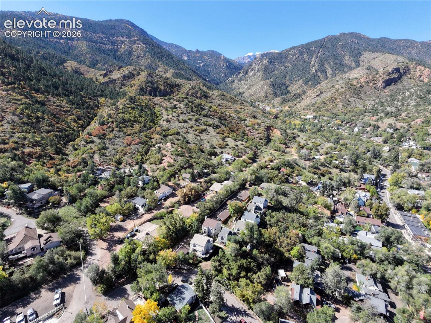 Mountainous terrain with natural vegetation, residential properties nestled among trees, and clear blue sky