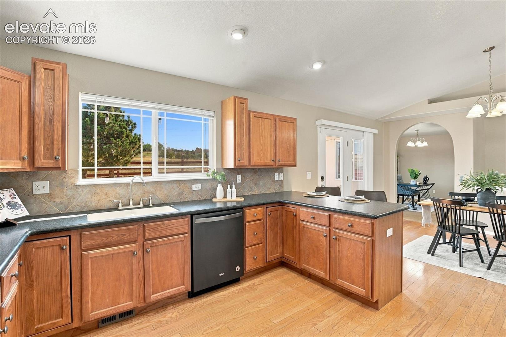 Wood-finish flooring, wood cabinetry, tile backsplash, black countertops, and a full-size dishwasher