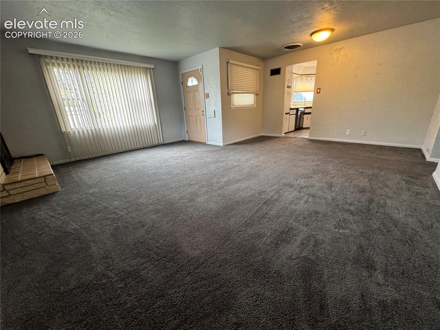 Open-concept interior featuring wall-to-wall carpeting, a floor-to-ceiling brick-surround fireplace, large windows with vertical blinds, and a solid-core entry door
