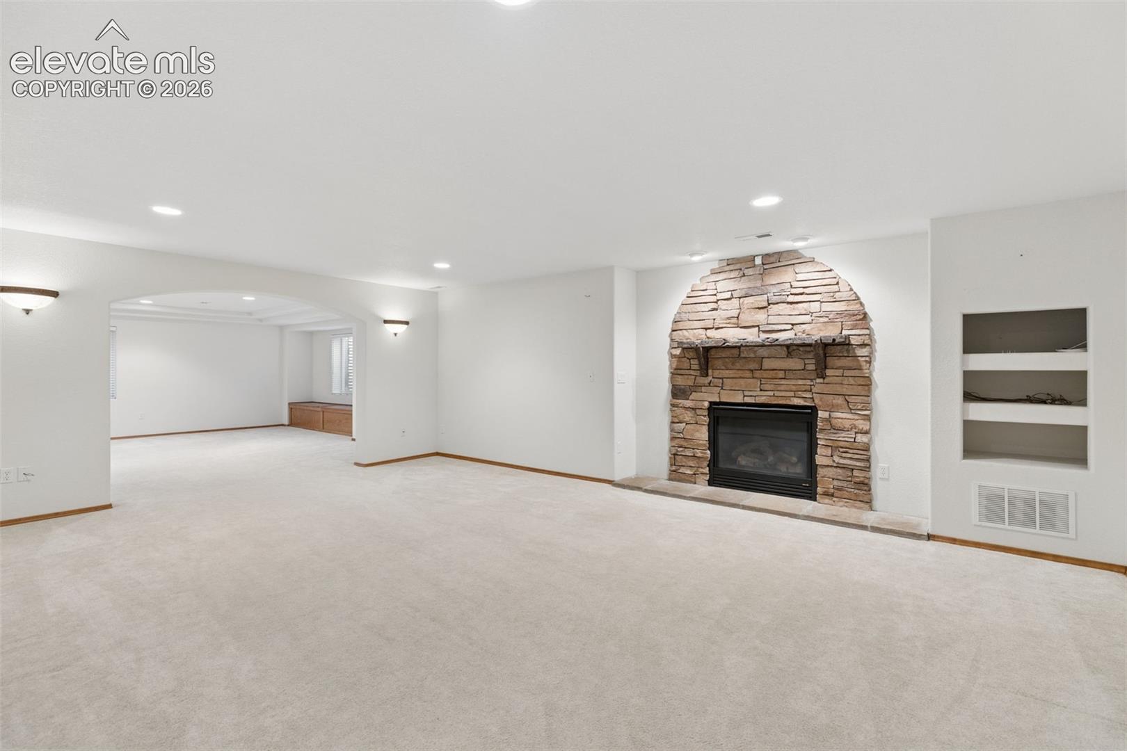 Spacious interior featuring a stone-clad fireplace with a wood mantel, recessed lighting, arched entryway, and built-in wall shelving