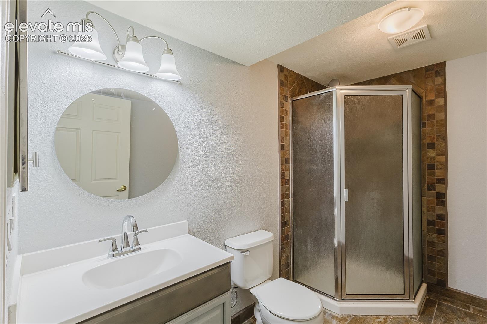 Basement bathroom featuring a circular framed mirror with integrated vanity lighting, with brushed nickel fixtures, corner shower enclosure with frameless glass doors and mosaic tile surround, and ceramic tile flooring