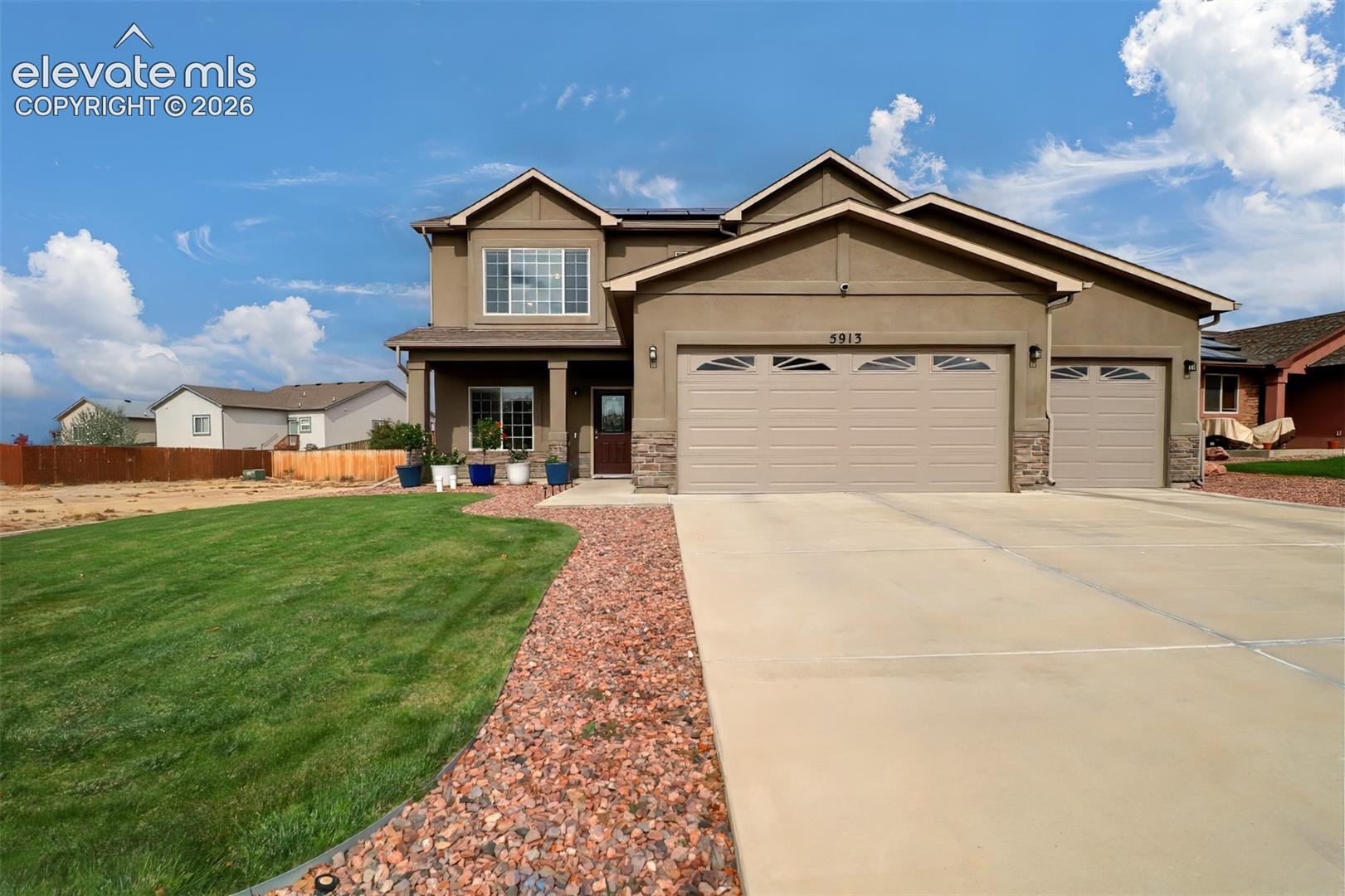 Two-story residence featuring a multi-tone stucco and stone facade, attached three-car garage, concrete driveway, and landscaped front lawn with stone-accented perimeter