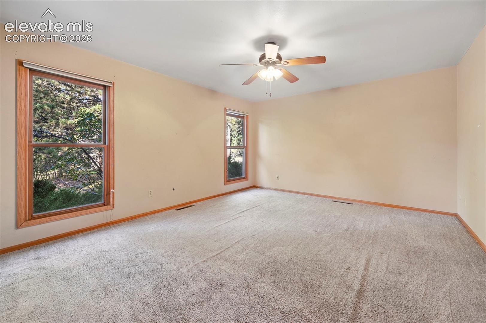 Two large windows with wood-finish trim, ceiling fan with integrated lighting, and neutral carpeting