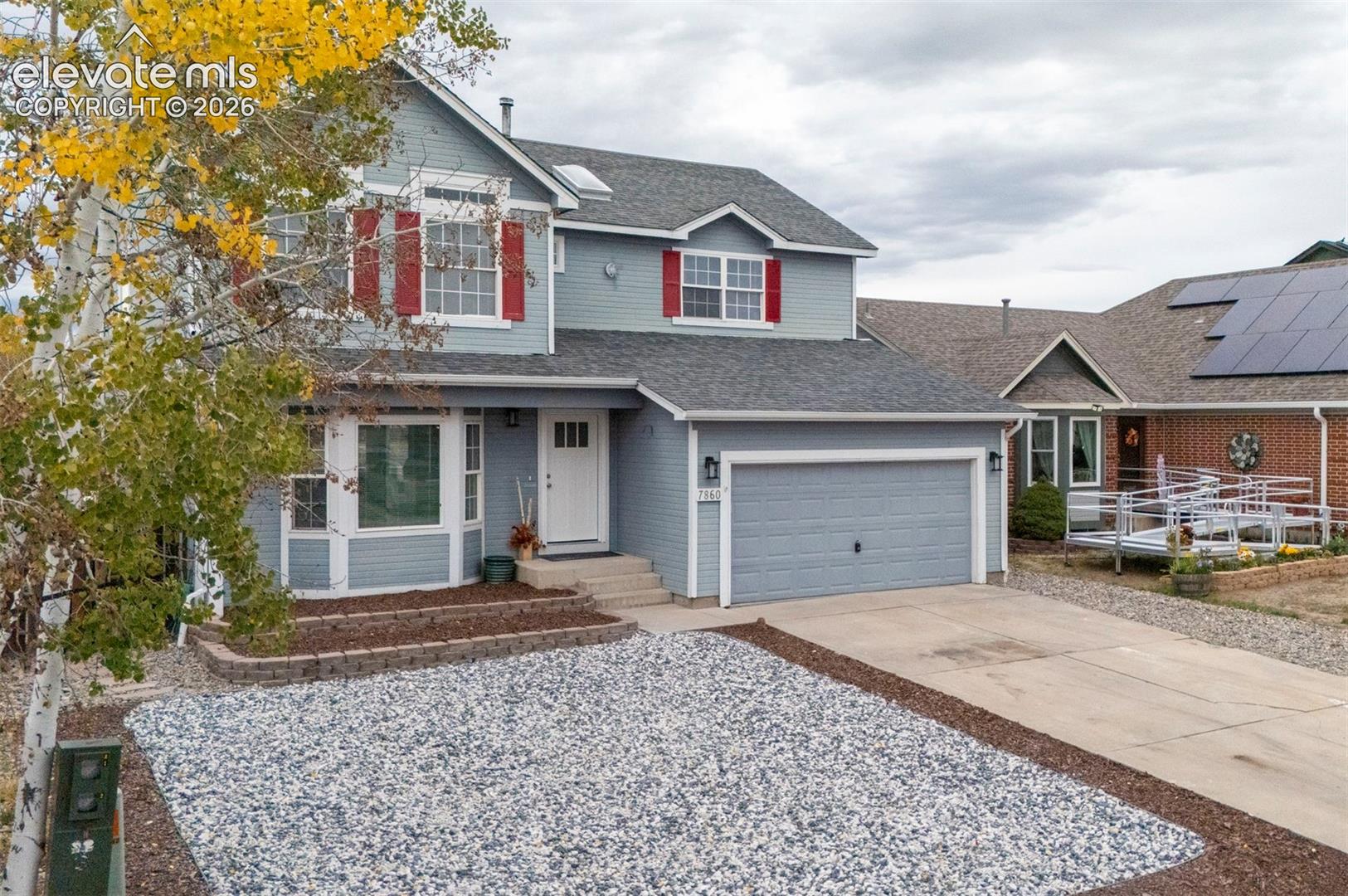 Two-story residence with lap siding, red window shutters, attached two-car garage, multi-gabled roofline, and front landscaping featuring decorative stone ground cover