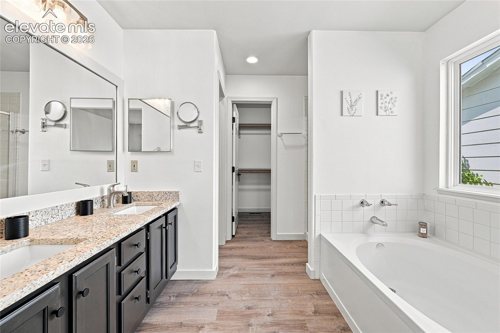 Dual vanity with granite-style countertops, wood-finish flooring, built-in bathtub, and an open closet with shelving