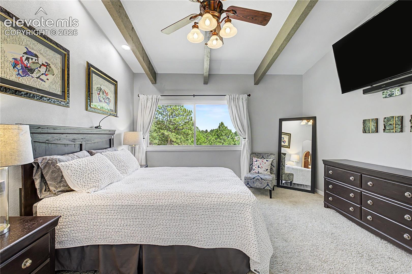 The primary bedroom continues the home's architectural theme with vaulted, beamed ceilings and a large window framing the wooded lot beyond.
