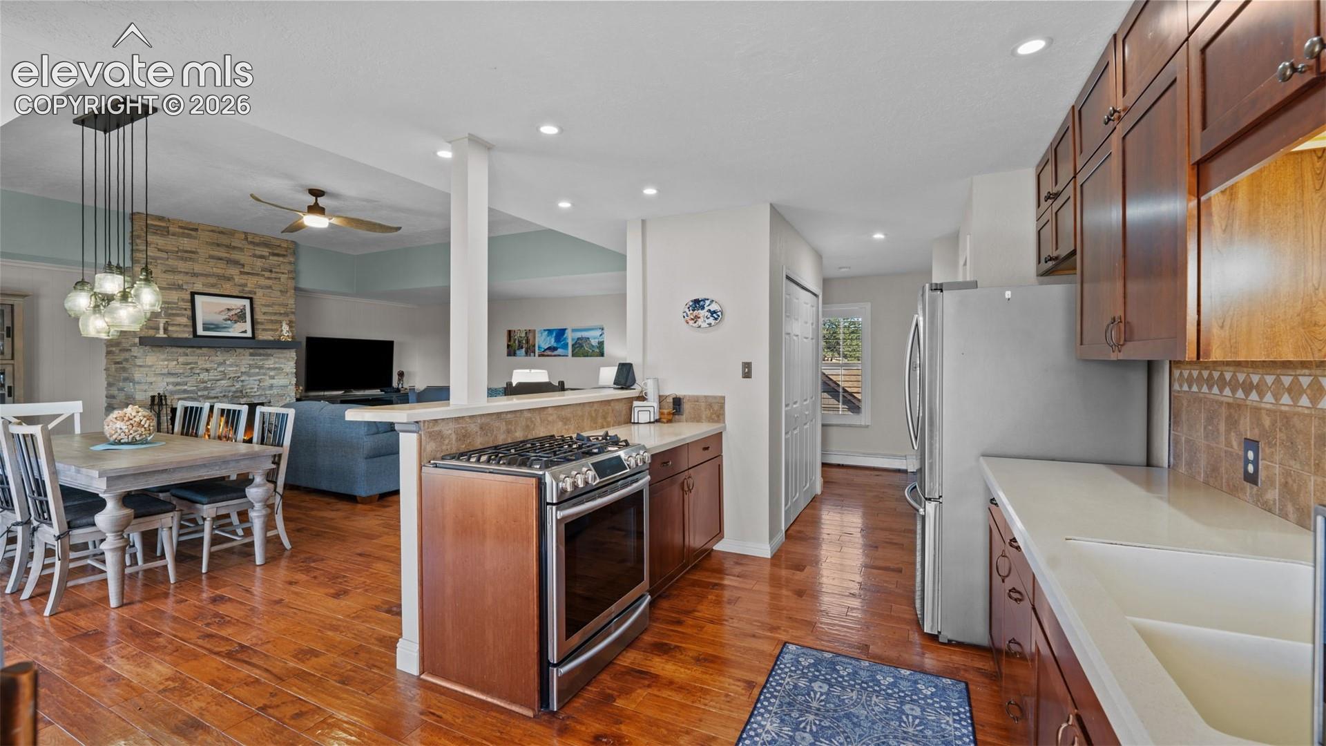 Kitchen featuring wood cabinetry, stainless steel gas range, stone-tiled backsplash, and composite countertops, with an attached dining area showcasing a multi-globe pendant light fixture and a stone fireplace