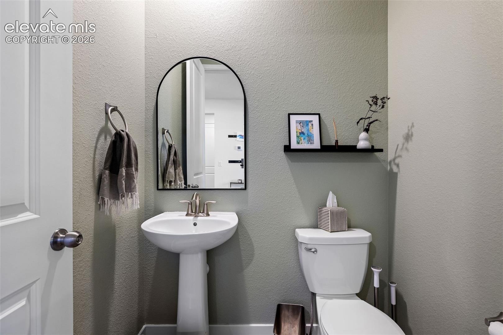 Pedestal sink with brushed nickel faucet, arched mirror, white toilet, and wall-mounted shelving
