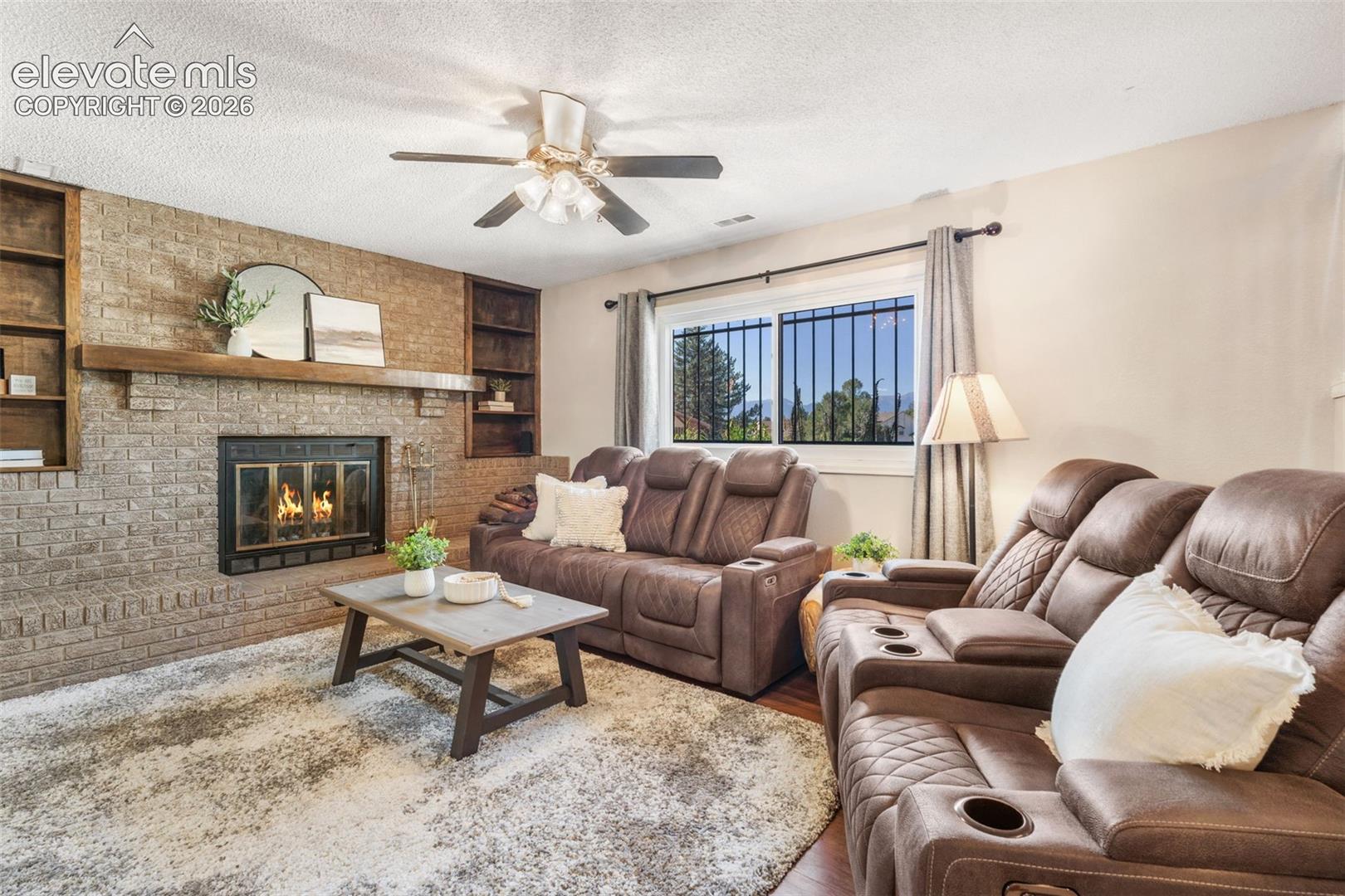 Full-height masonry fireplace with wood mantel, integrated wood shelving, and ceiling fan with light kit