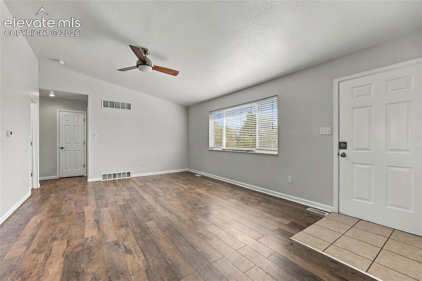 Living area featuring vaulted ceilings, wood-finish flooring, a ceiling fan, a window with horizontal blinds, and a tiled entryway