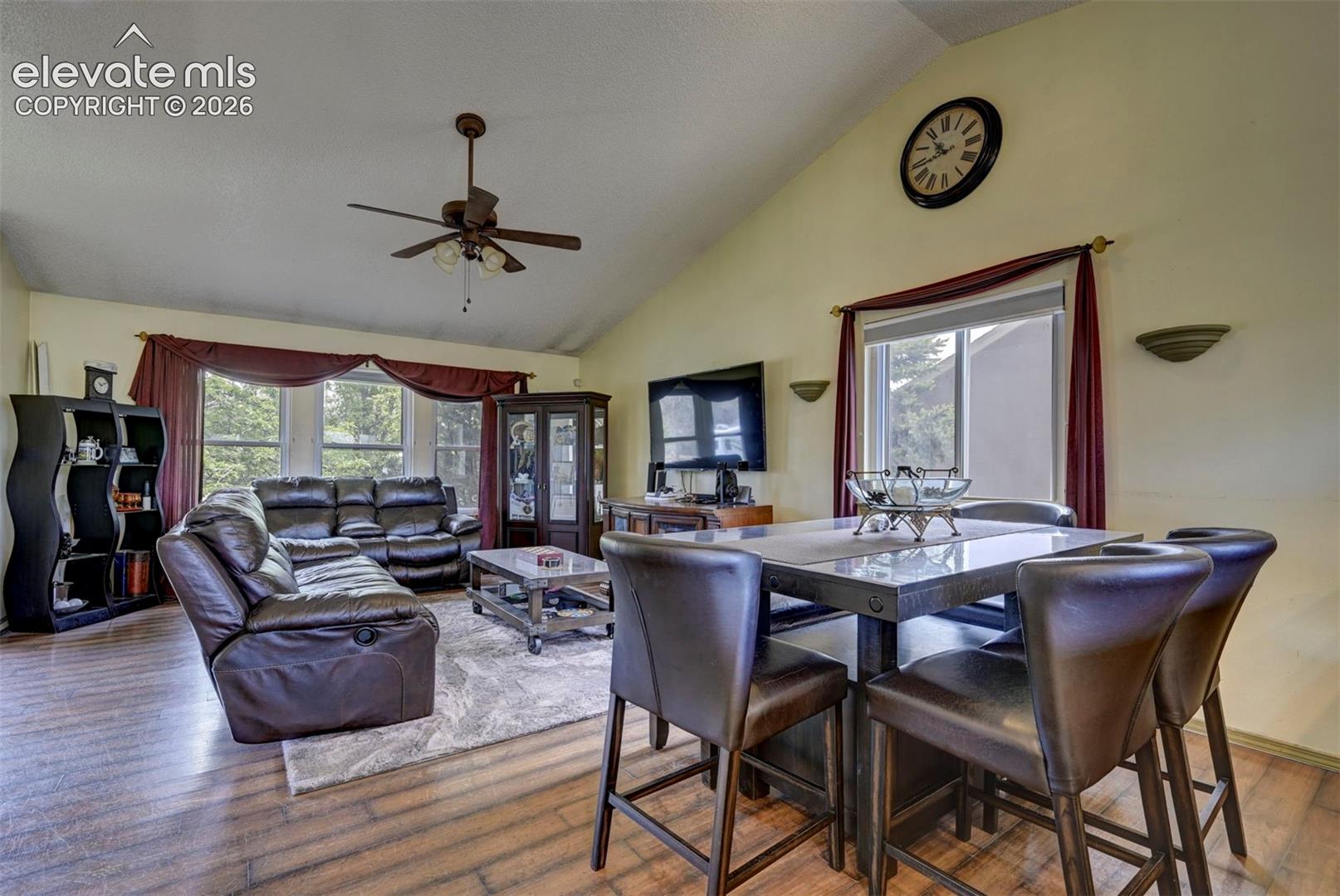 Open-concept living area featuring vaulted ceilings, wood-finish flooring, a ceiling fan with integrated lighting, and multiple windows