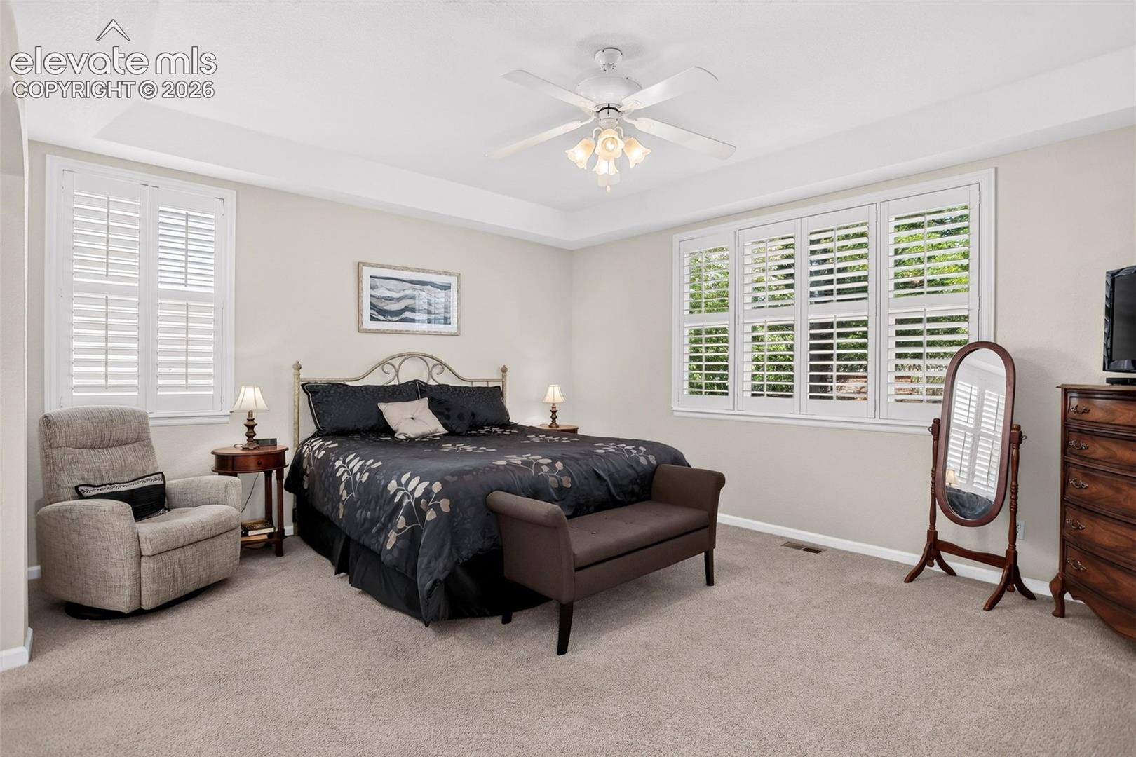 Tray ceiling, overhead fan with integrated lighting, plantation shutters, wall-to-wall carpeting, and neutral wall tones
