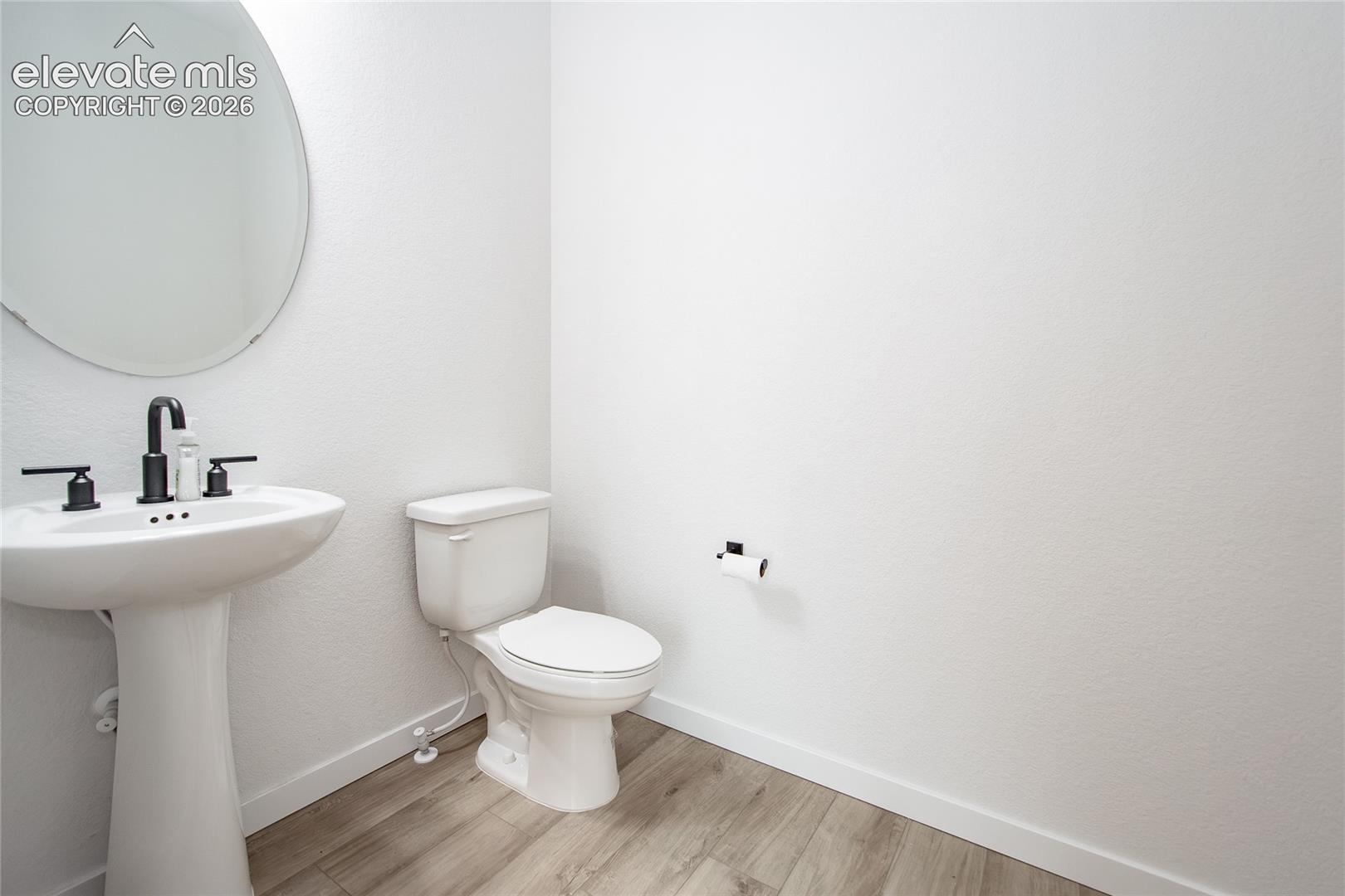 Pedestal sink with matte black widespread faucet, oval wall-mounted mirror, wood-finish flooring, and matching matte black hardware