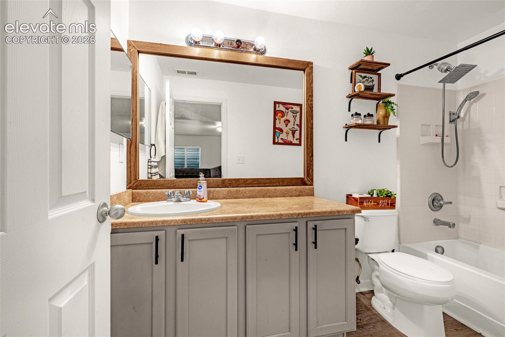 Bathroom featuring a wood-framed mirror, vanity with a light-colored countertop, gray cabinetry with black hardware, white subway tile shower surround, and wood-finish flooring