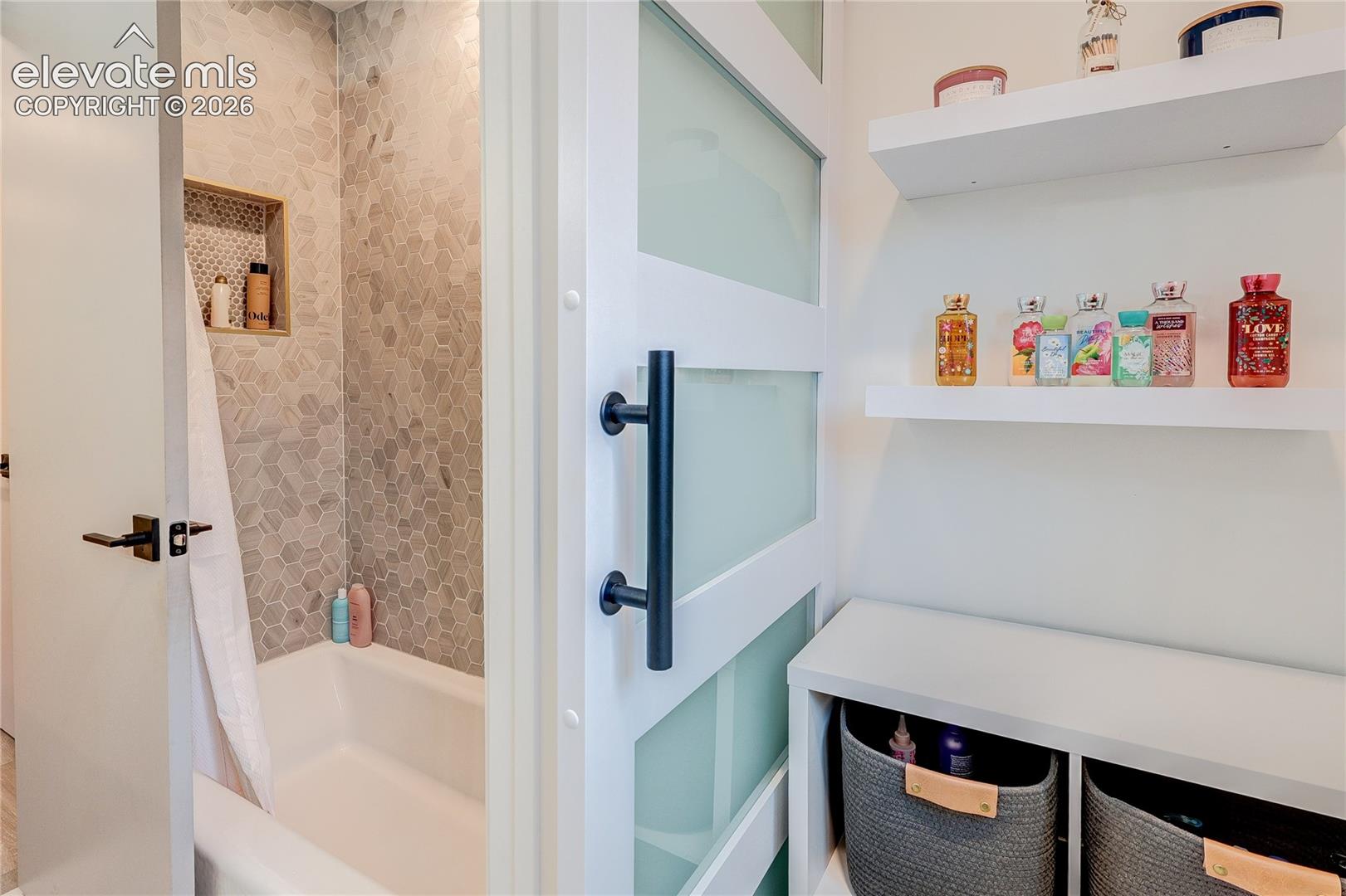 Sliding barn door with frosted panels, white floating shelves, built-in shelving unit, and a bathtub with hexagonal wall tile surround