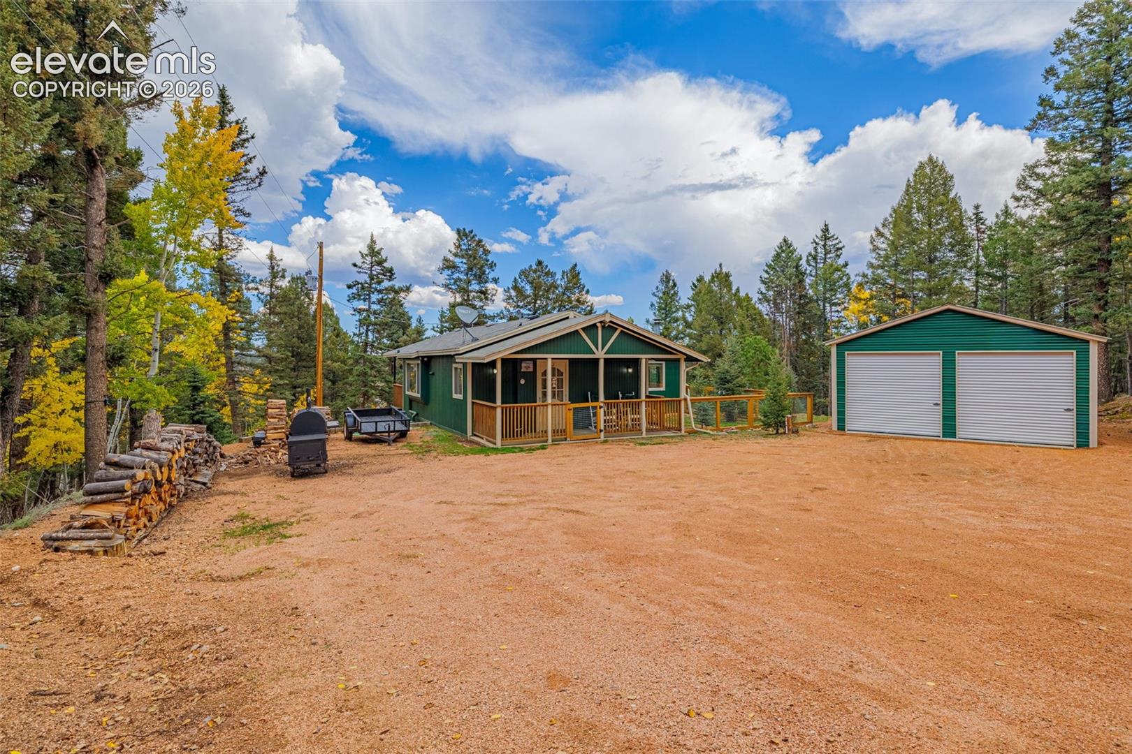 Single-level cabin exterior featuring green wood siding, a covered front porch with wooden railings, and a detached two-car garage with white overhead doors, all situated on a gravel lot