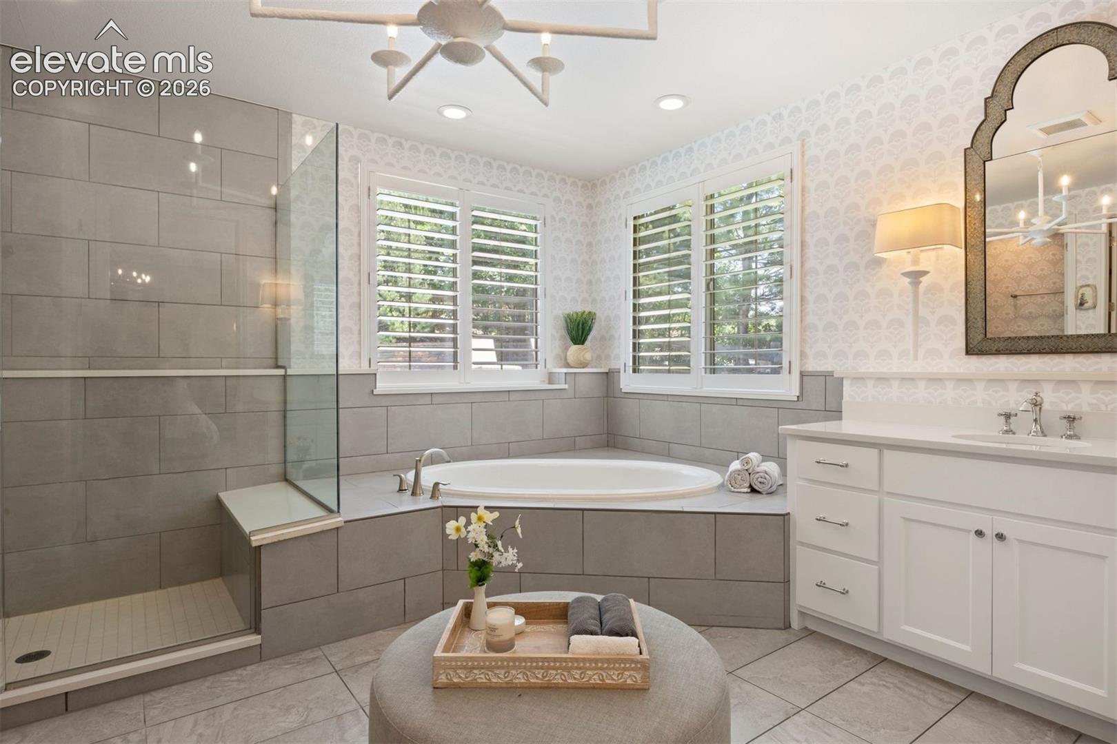Spacious bathroom featuring a built-in soaking tub, a walk-in shower with a glass enclosure and built-in bench, a single vanity with a quartz countertop, and plantation shutters