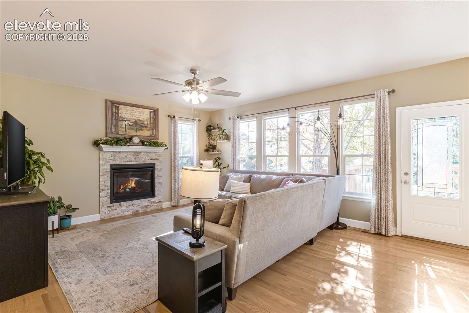 Living area featuring wood-finish flooring, a stone-surround fireplace with a mantel, a ceiling fan with integrated lighting, and multiple windows with exterior exposure