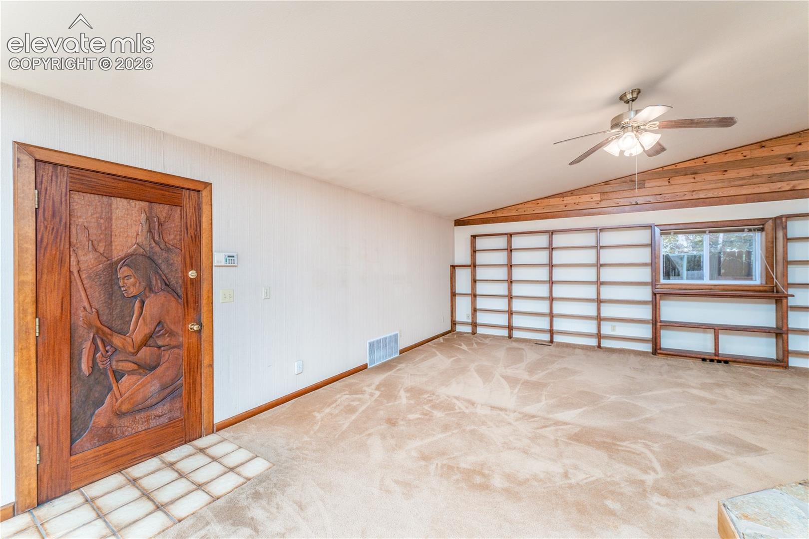 Vaulted ceiling with wood paneling, ceiling fan, expansive built-in shelving, decorative carved wood entry door, and tiled entry