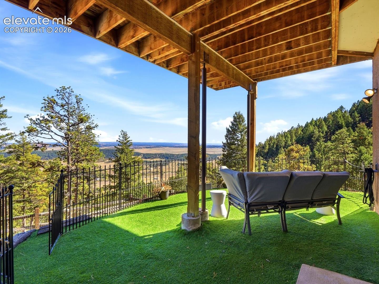 Walk out to Covered patio featuring an exposed wood beam ceiling, synthetic turf surfacing, metal perimeter fencing, and prominent forest scenery