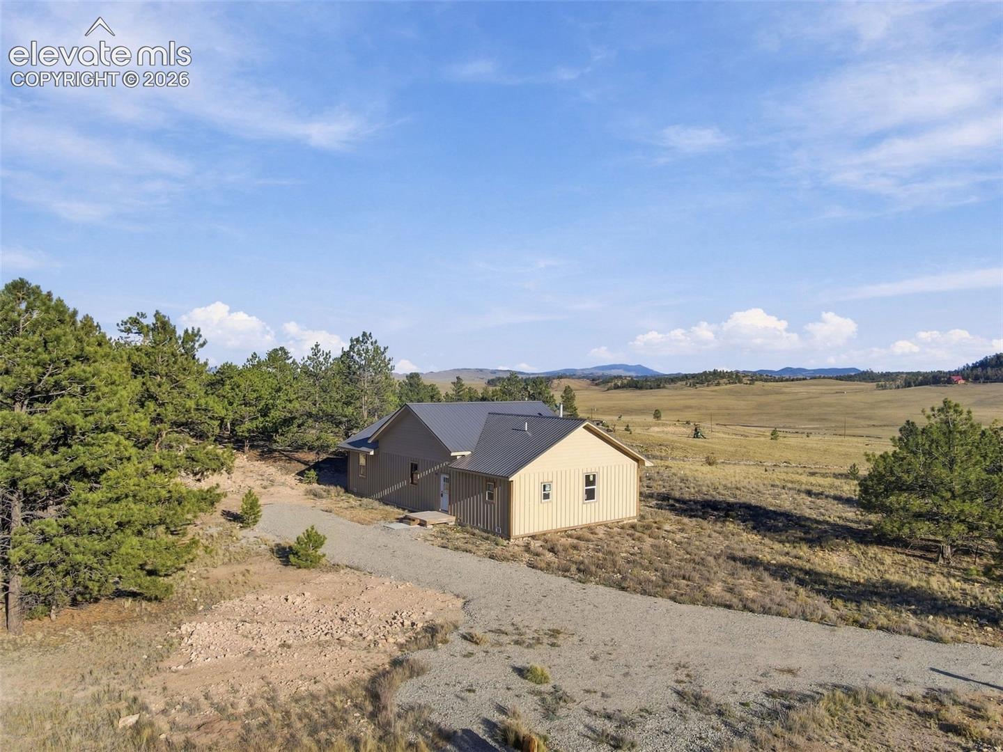 Single-story residence featuring vertical siding, a metal roof, and a gravel driveway set amidst a landscape of mature pines and open prairie terrain