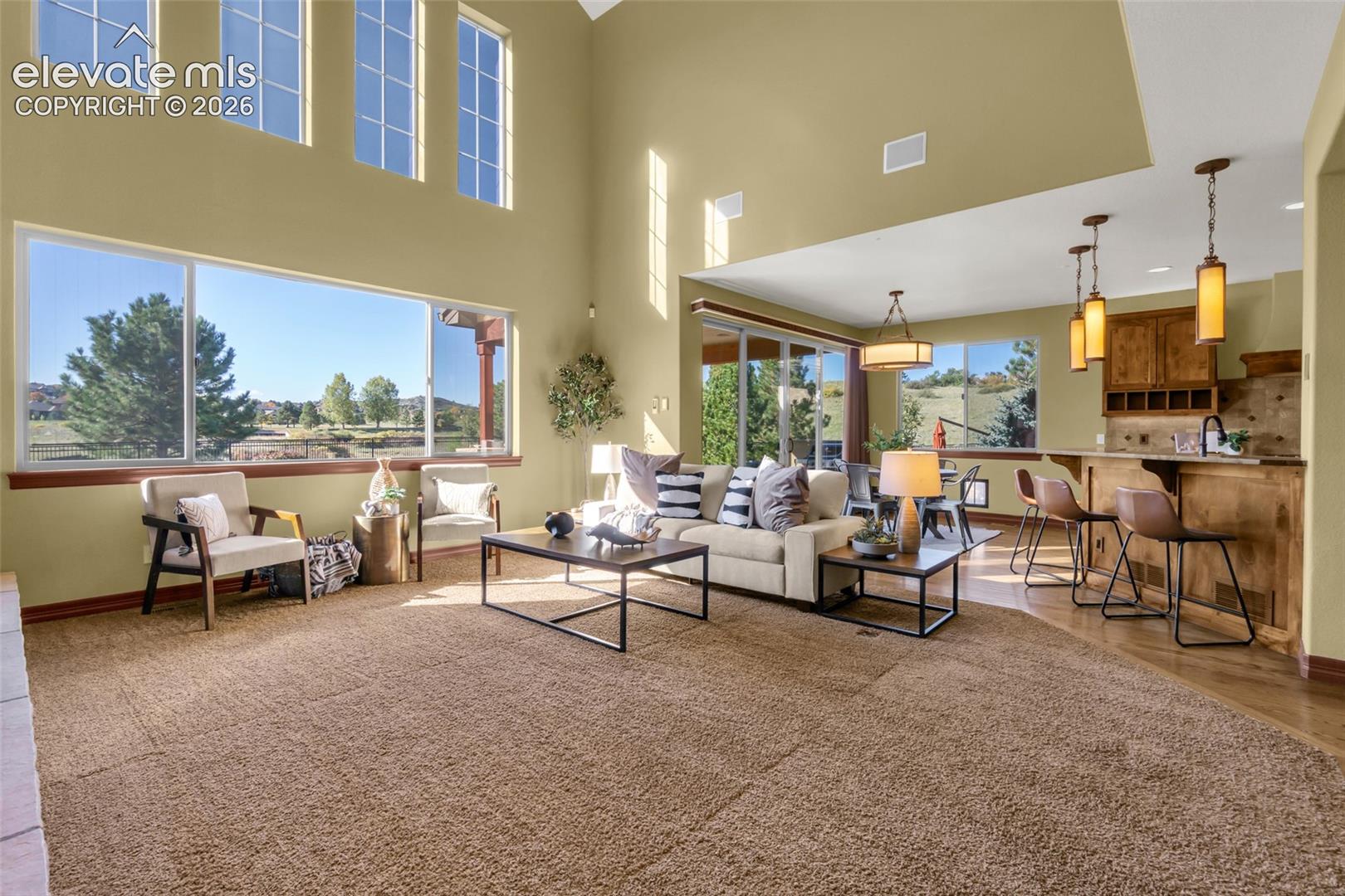 Expansive open-concept living area featuring vaulted ceilings, large multi-pane windows, a kitchen peninsula with wood-finish cabinetry, and decorative pendant lighting