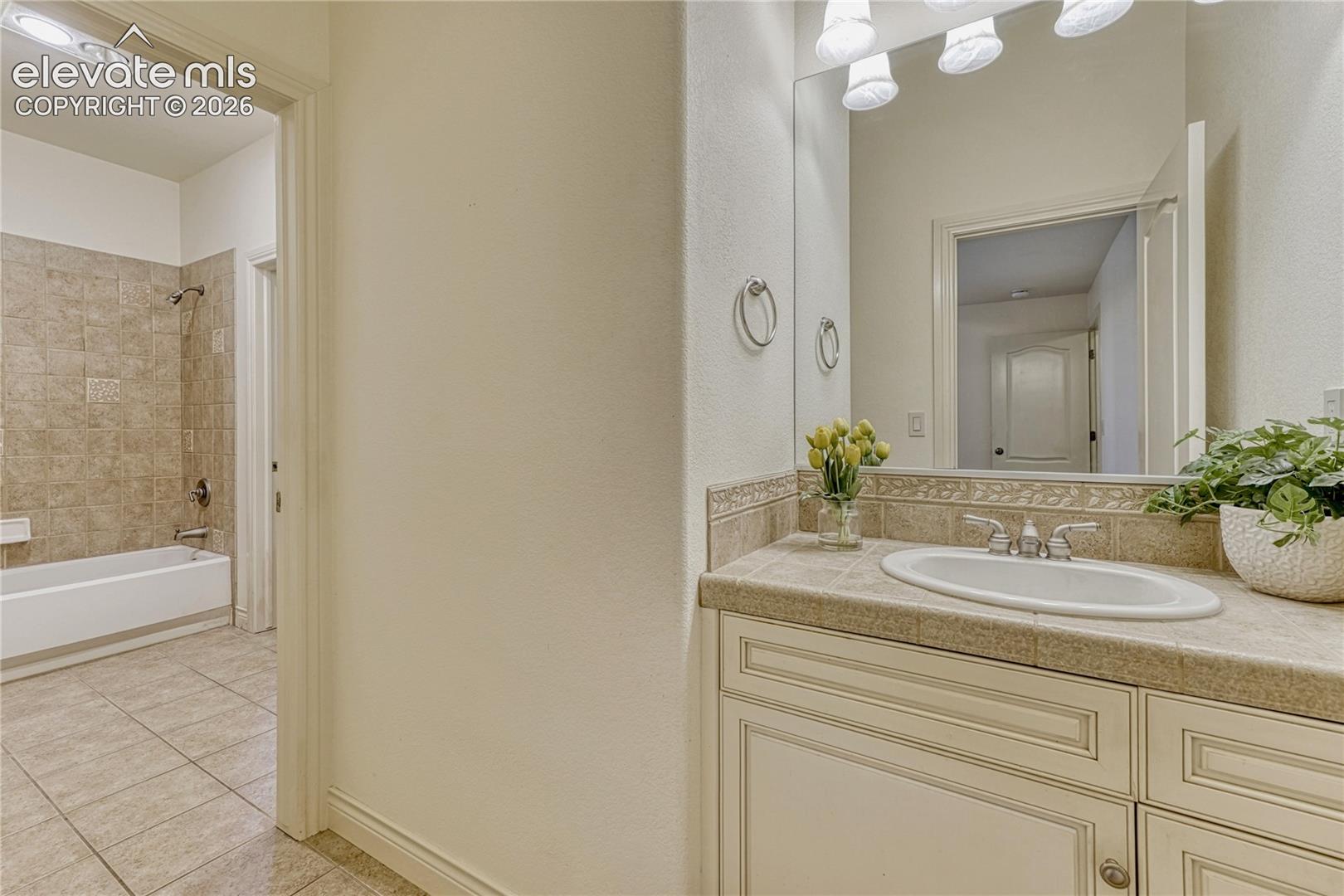 Vanity featuring light-tone cabinetry, tiled countertop with decorative backsplash, and inset sink, accompanied by a tub and shower combination with full-height tile surround and large-format tile flooring