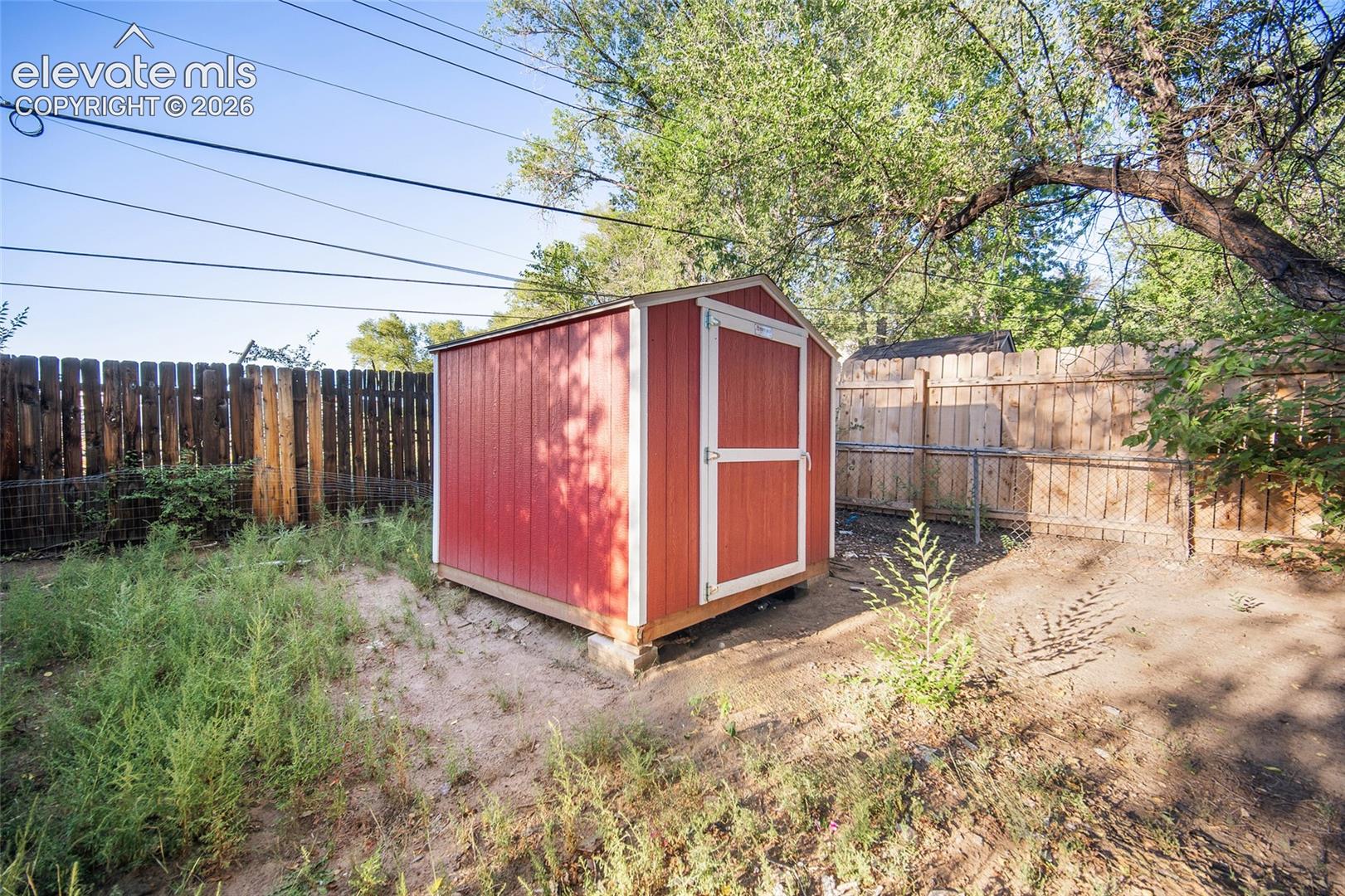 Red storage shed with white trim, wood fence, and mature tree