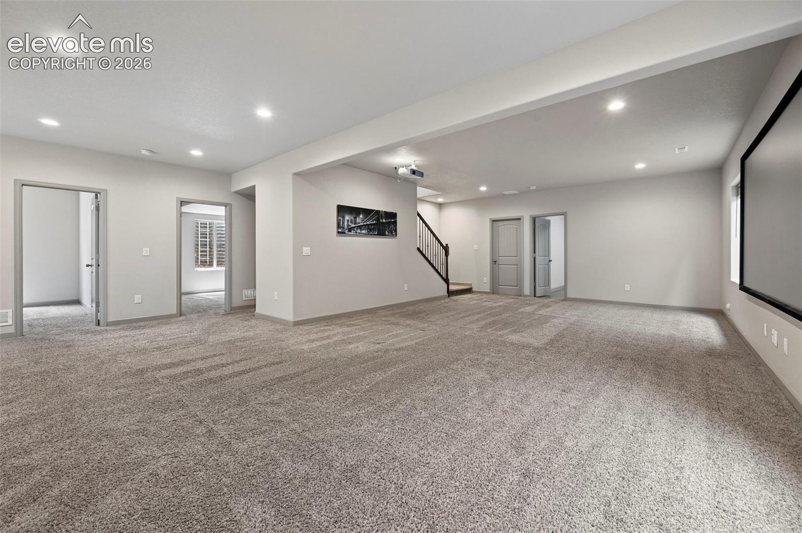 Open-concept space featuring recessed ceiling lighting, wall-to-wall carpeting, a large wall-mounted projection screen, and a staircase with dark wood-finish handrails