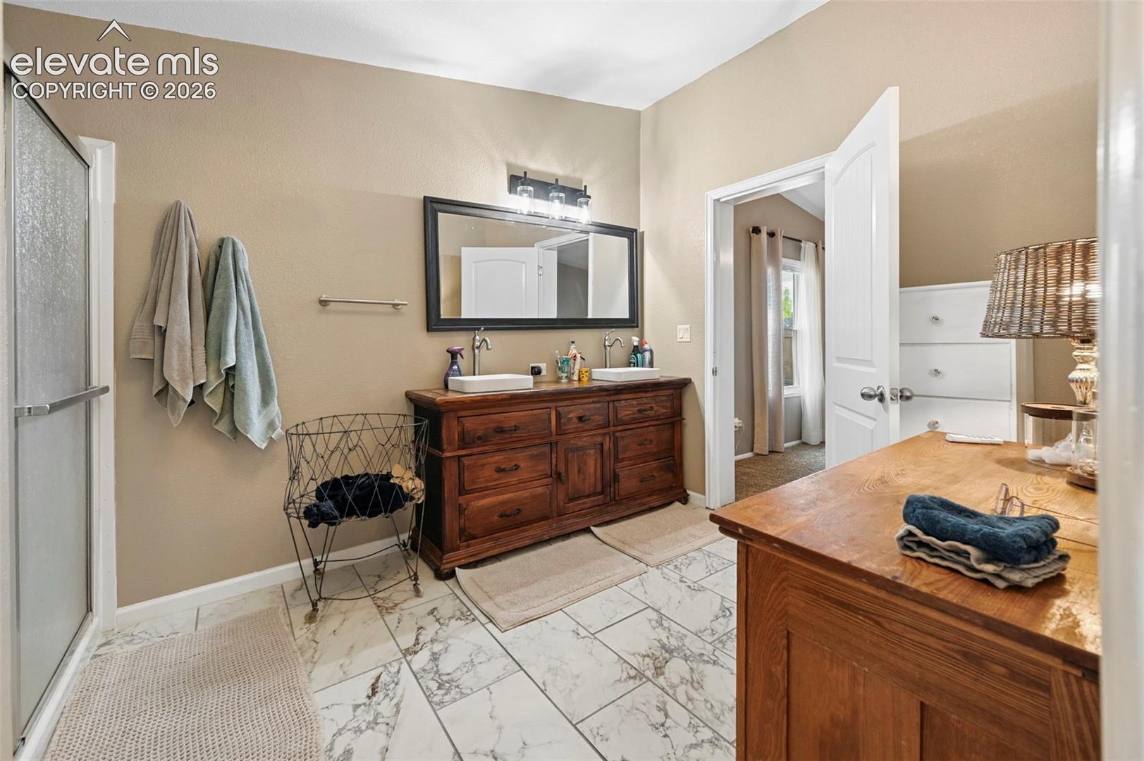 Double vanity featuring a stained-wood cabinet, dual vessel sinks, and a large framed mirror with three-bulb overhead lighting, complemented by patterned tile flooring and a glass-enclosed shower
