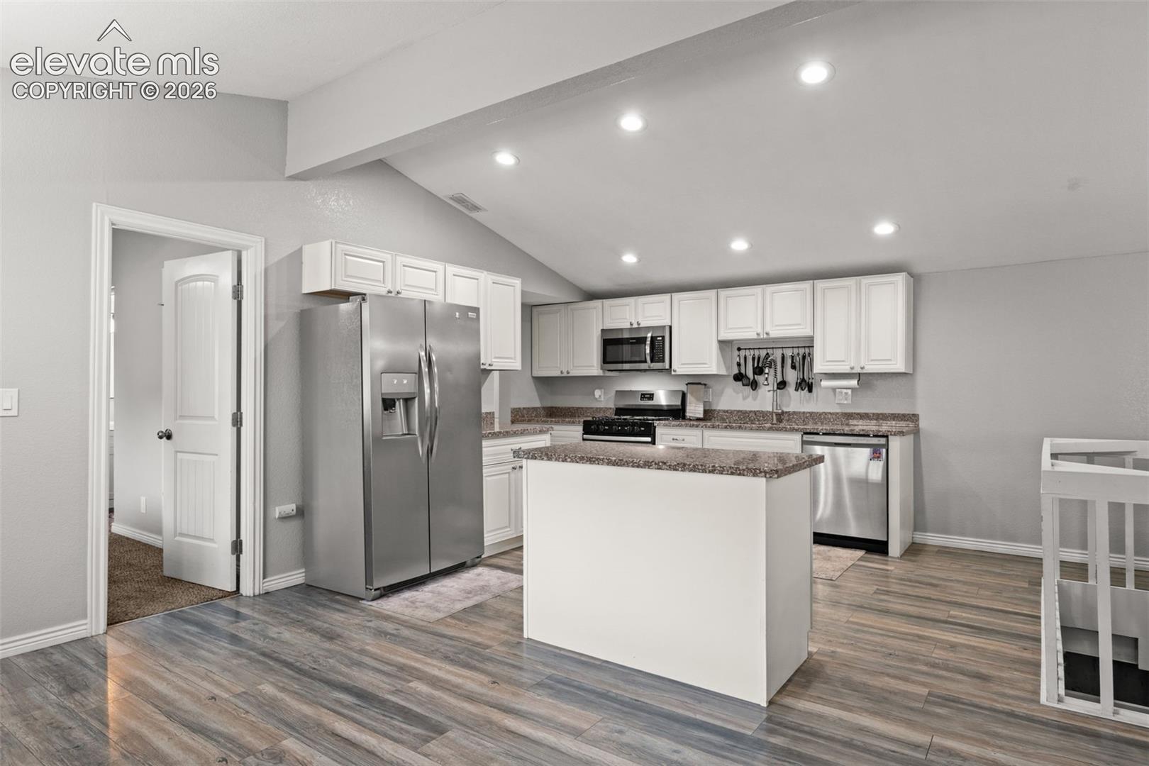 Kitchen featuring vaulted ceilings, wood-finish flooring, white shaker-style cabinetry, stone-finish countertops, and stainless steel appliances