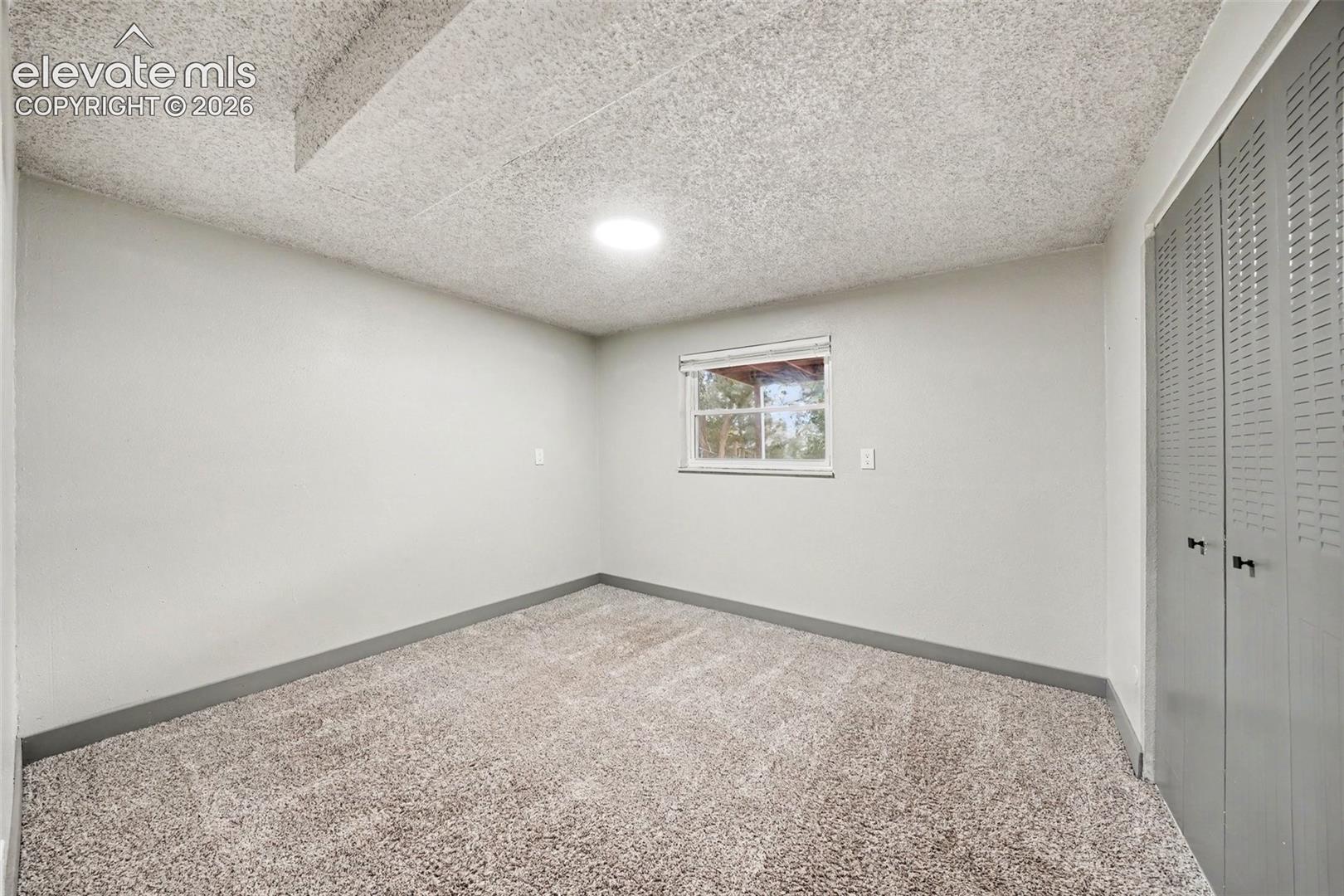 Room featuring neutral-toned carpeting, textured ceiling, double-door closet with louvered panels, and a single-pane window
