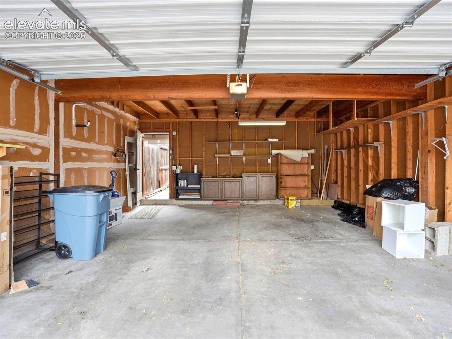 Attached two-car garage featuring exposed wood framing, concrete flooring, built-in wooden cabinetry, integrated wall shelving, and a metal roll-up door