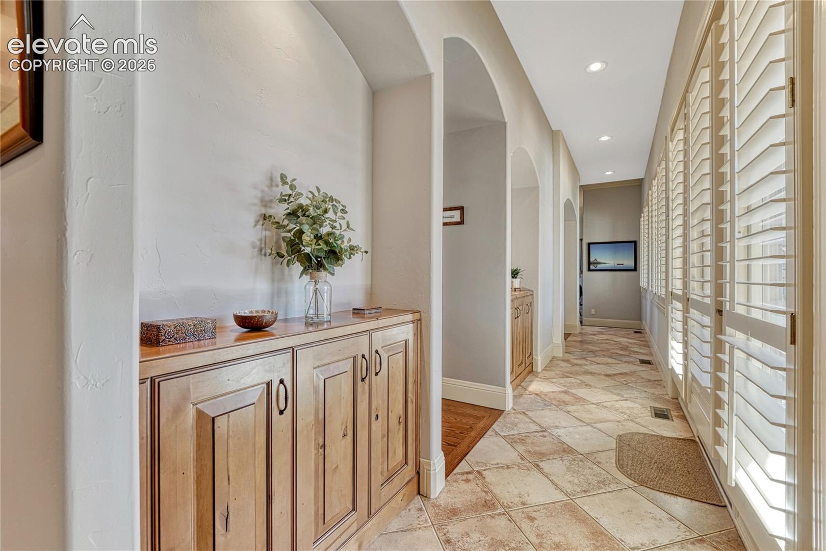 Hallway featuring arched architectural openings, built-in cabinetry with natural wood finish, stone-tile flooring, and plantation shutters