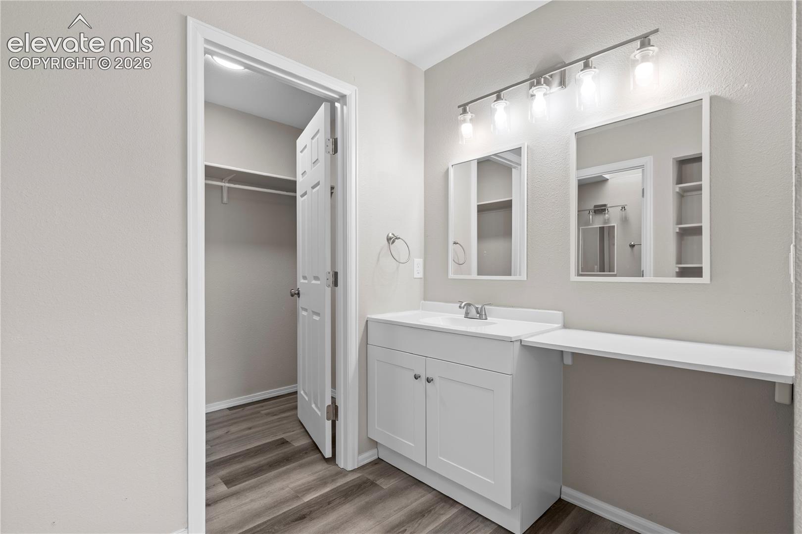 Vanity featuring a white cabinet base, integrated countertop and basin, dual wall-mounted mirrors, overhead light fixture, and an extended shelf, complemented by wood-finish flooring and a walk-in closet