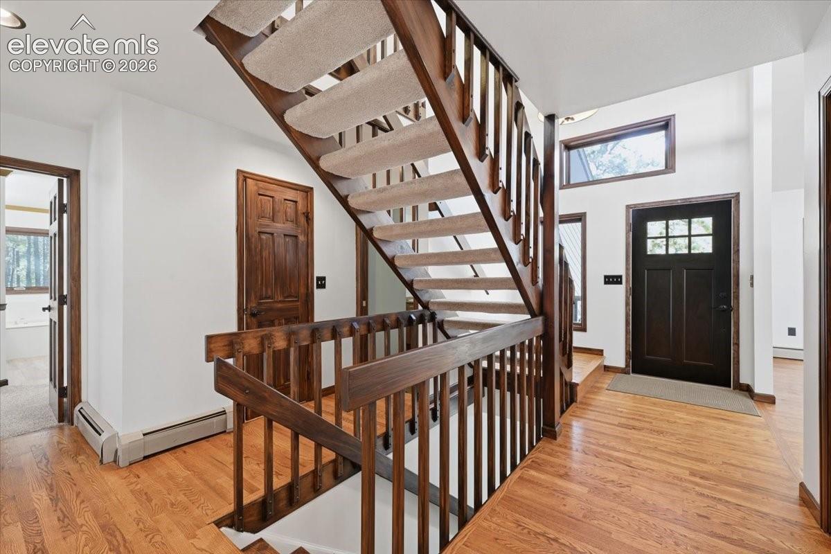 Hardwood flooring, wood staircase with carpeted treads, dark wood entry door with glass panes, wood trim around windows, and white walls