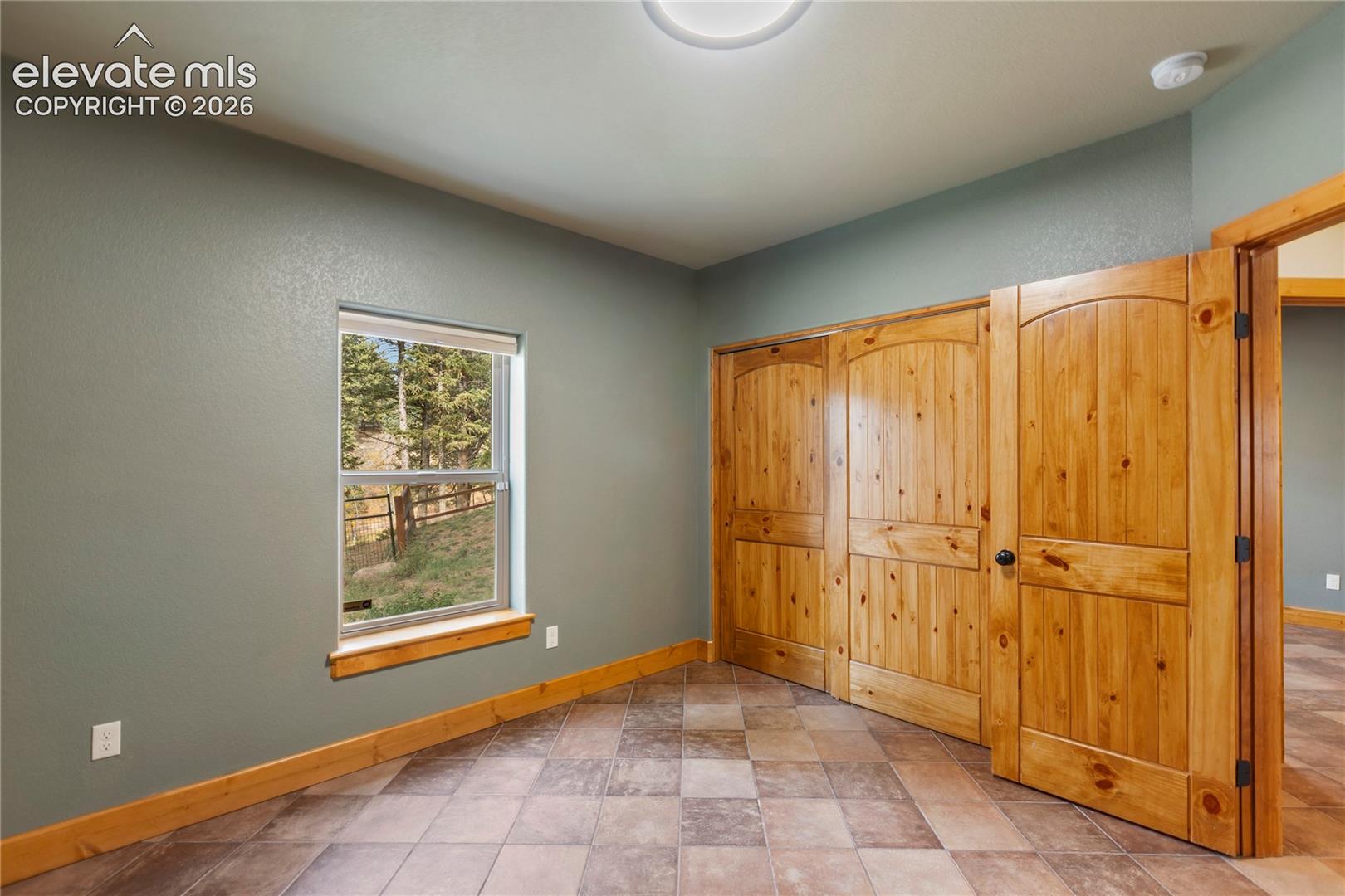 Room featuring tiled flooring, natural wood-finish interior doors with arched detailing, matching wood window trim, and a window framing exterior woodland scenery