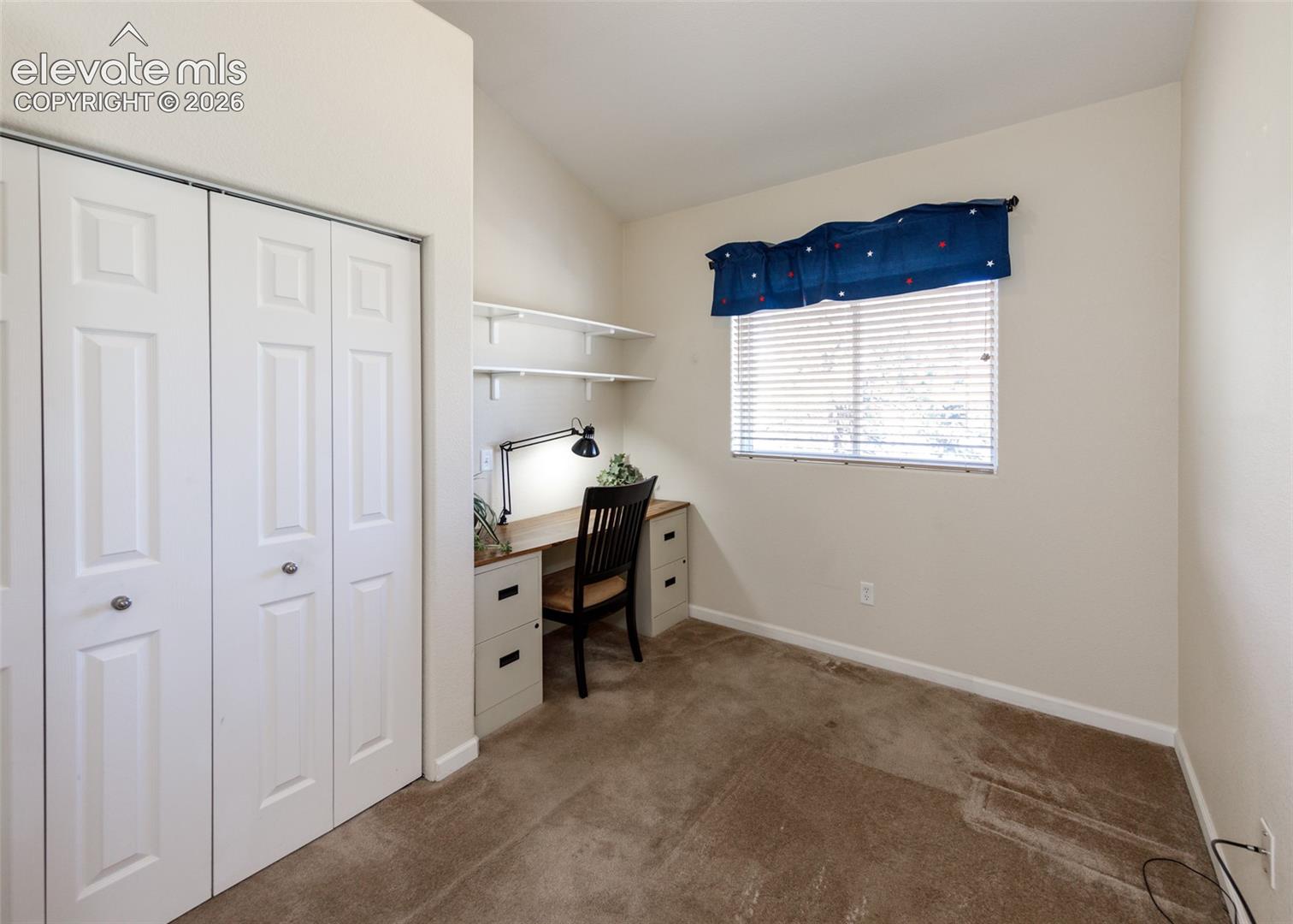 Room featuring a vaulted ceiling, neutral wall coloring, carpeted flooring, bi-fold closet doors, and built-in desk cabinetry with integrated shelving