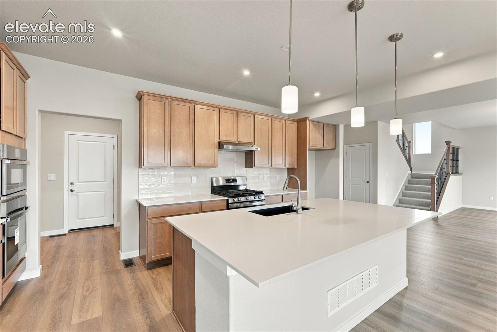 Kitchen featuring wood cabinetry, quartz countertops, a central island with an undermount sink, built-in stainless steel appliances, and pendant lighting