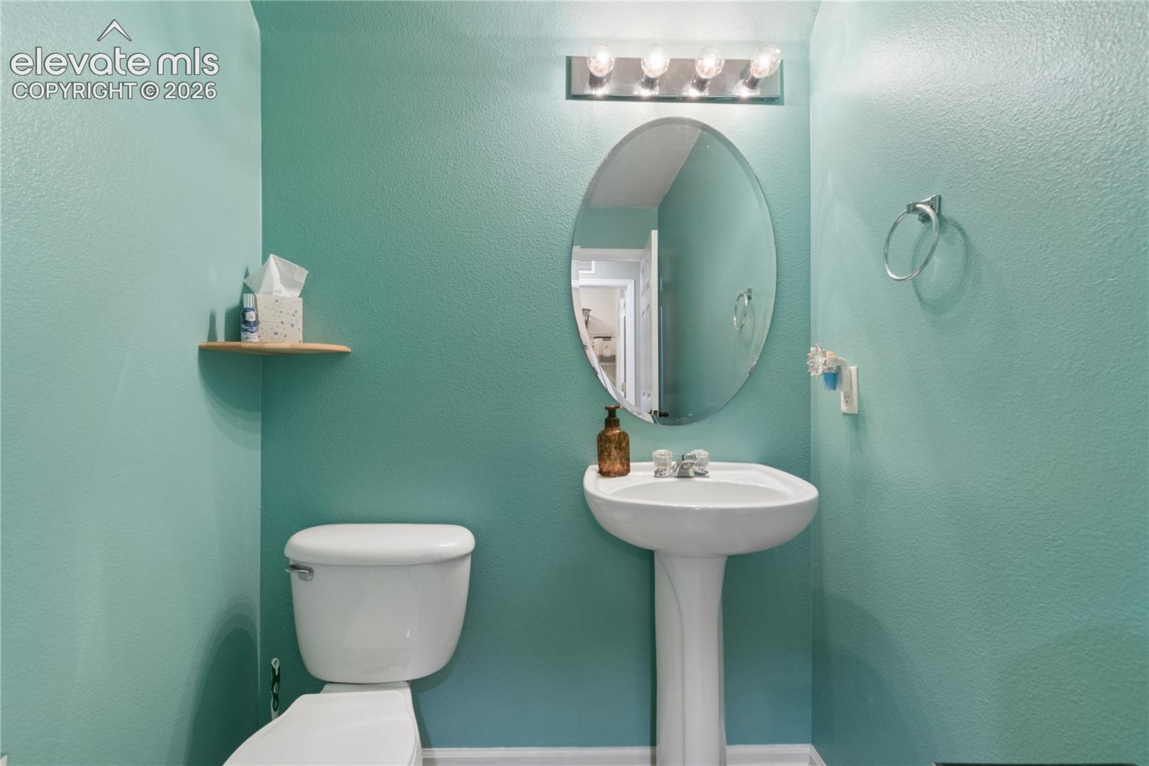 Pedestal sink with an oval mirror, above a toilet with a wall-mounted floating shelf, all illuminated by a four-bulb vanity light