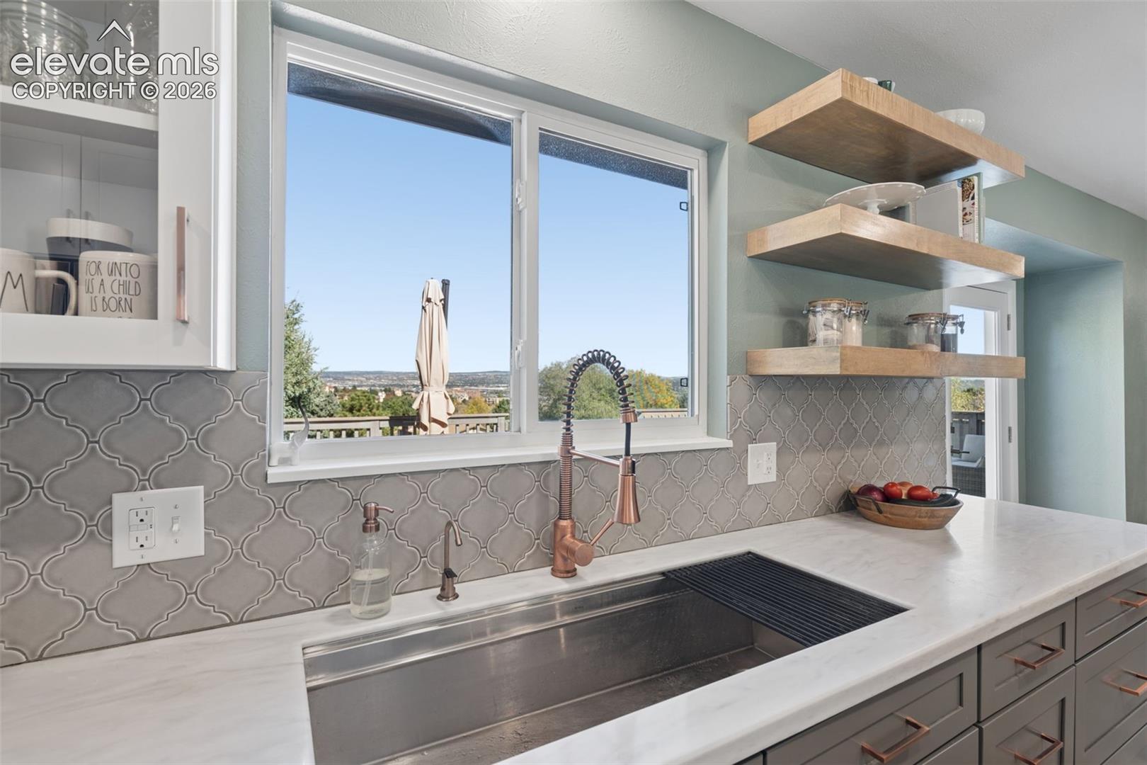 Kitchen featuring a stainless steel farmhouse-style sink, copper-toned gooseneck faucet, light-toned stone-finish countertops, grey arabesque tile backsplash, and floating wood-finish shelving
