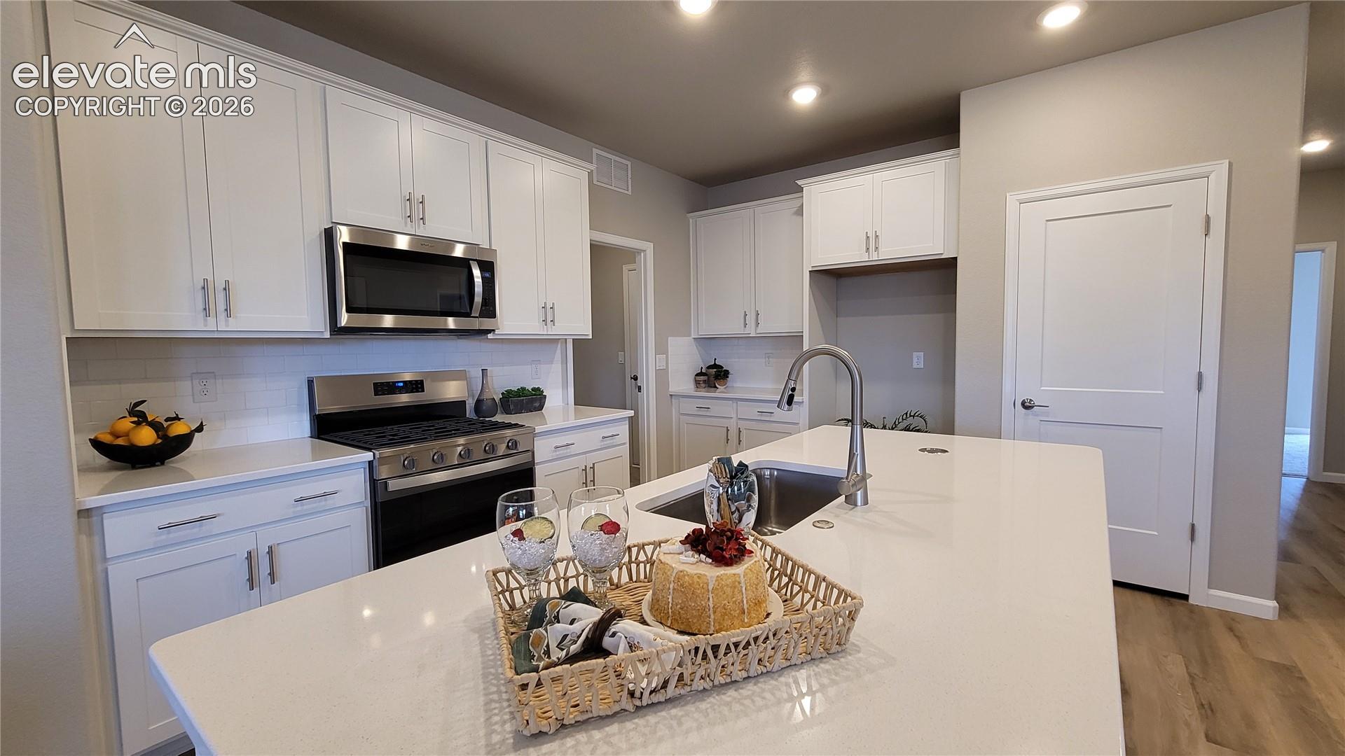 Kitchen featuring white shaker-style cabinetry, quartz countertops, a central island with a stainless steel sink, LED lighting, and LVP flooring, pantry with wood shelving, and stainless steel gas range, microwave, and dishwasher!
